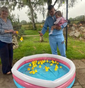 Family playing duck pond game at a summer fete. Young man holding baby, woman with fishing rod.