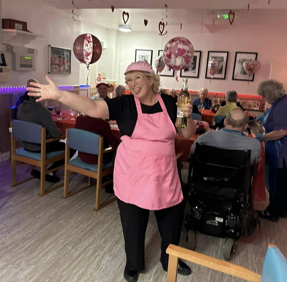 417476930_839844838156005_5713192493575131950_n Woman in pink apron welcomes guests to a Valentine's Day 2024 party.